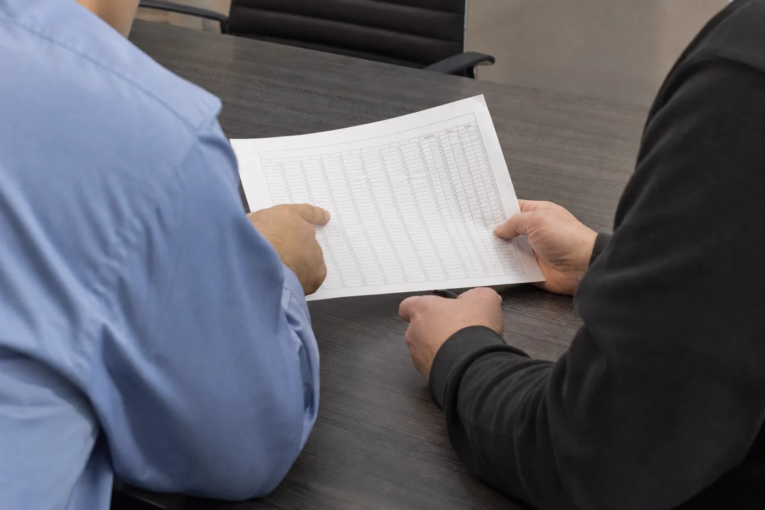 Two people at a dark wood conference table reviewing a printed page of figures in a table, one pointing to a row while the other holds the sheet and a pen