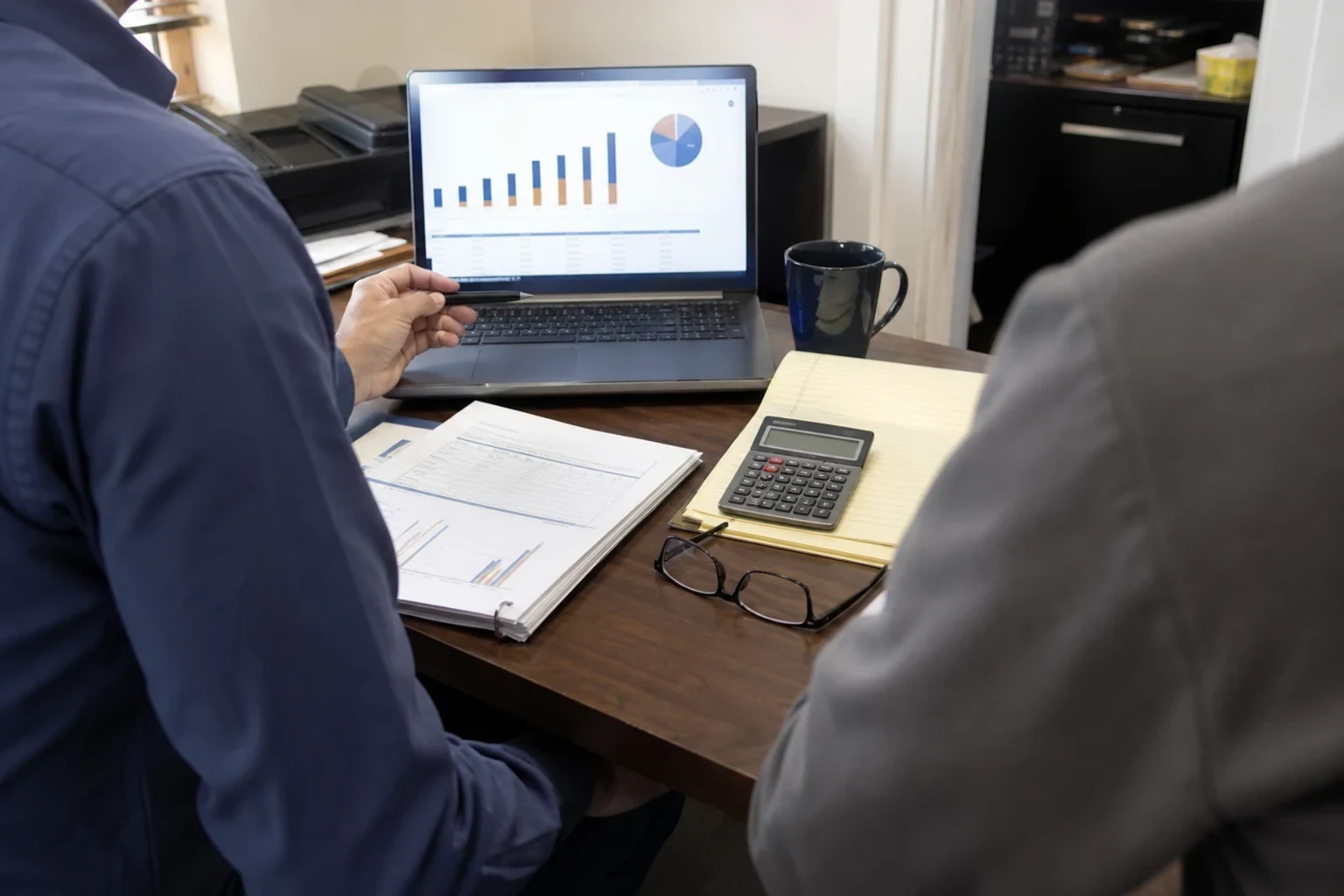 Over-the-shoulder view of two people at an office desk reviewing a laptop with bar and pie charts, a binder of financial reports, a calculator and a legal pad